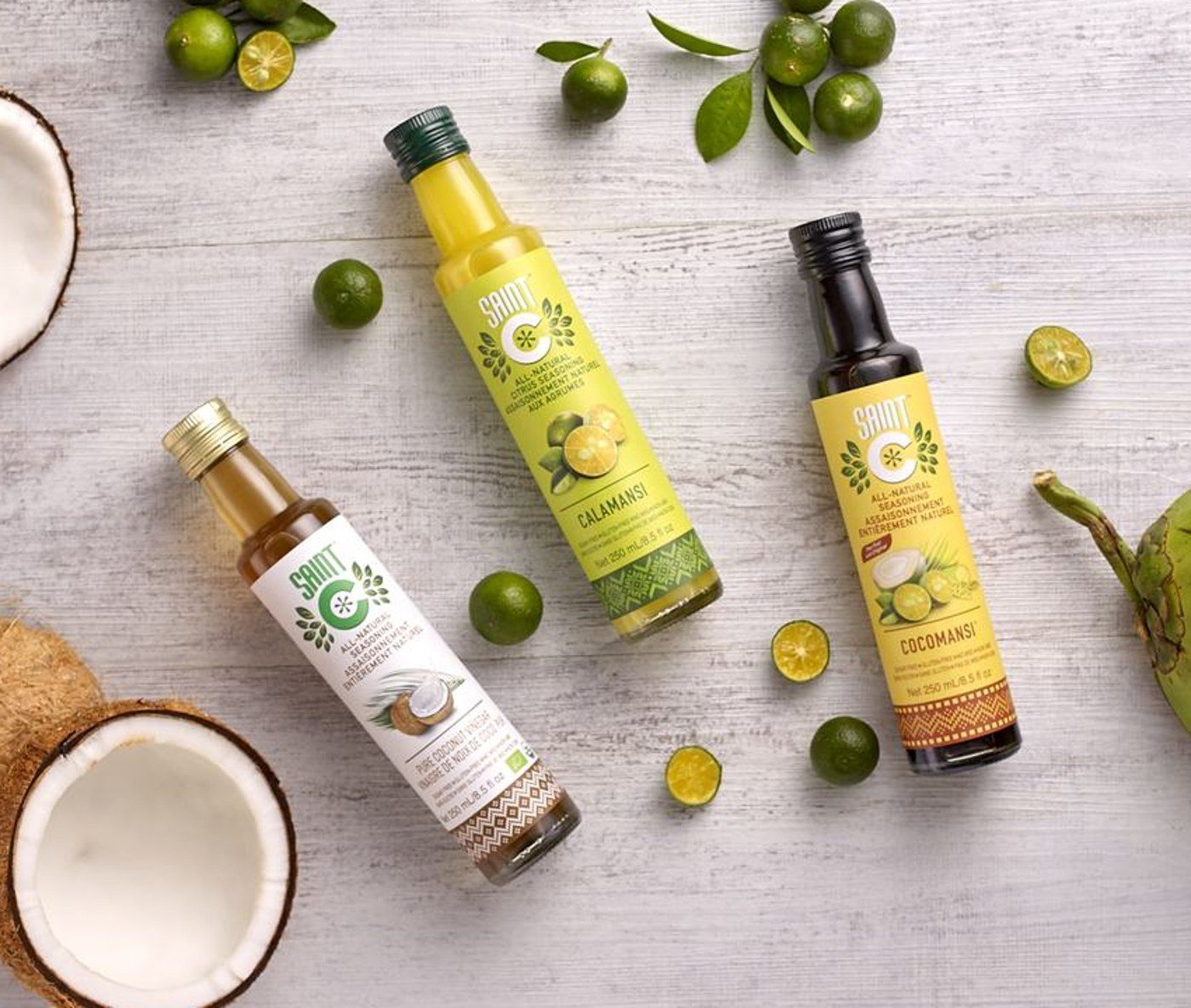 Saint C Pure Coconut Vinegar, Calamansi and Cocomansi bottles lying among fresh coconuts and calamansi limes on a pale wooden table